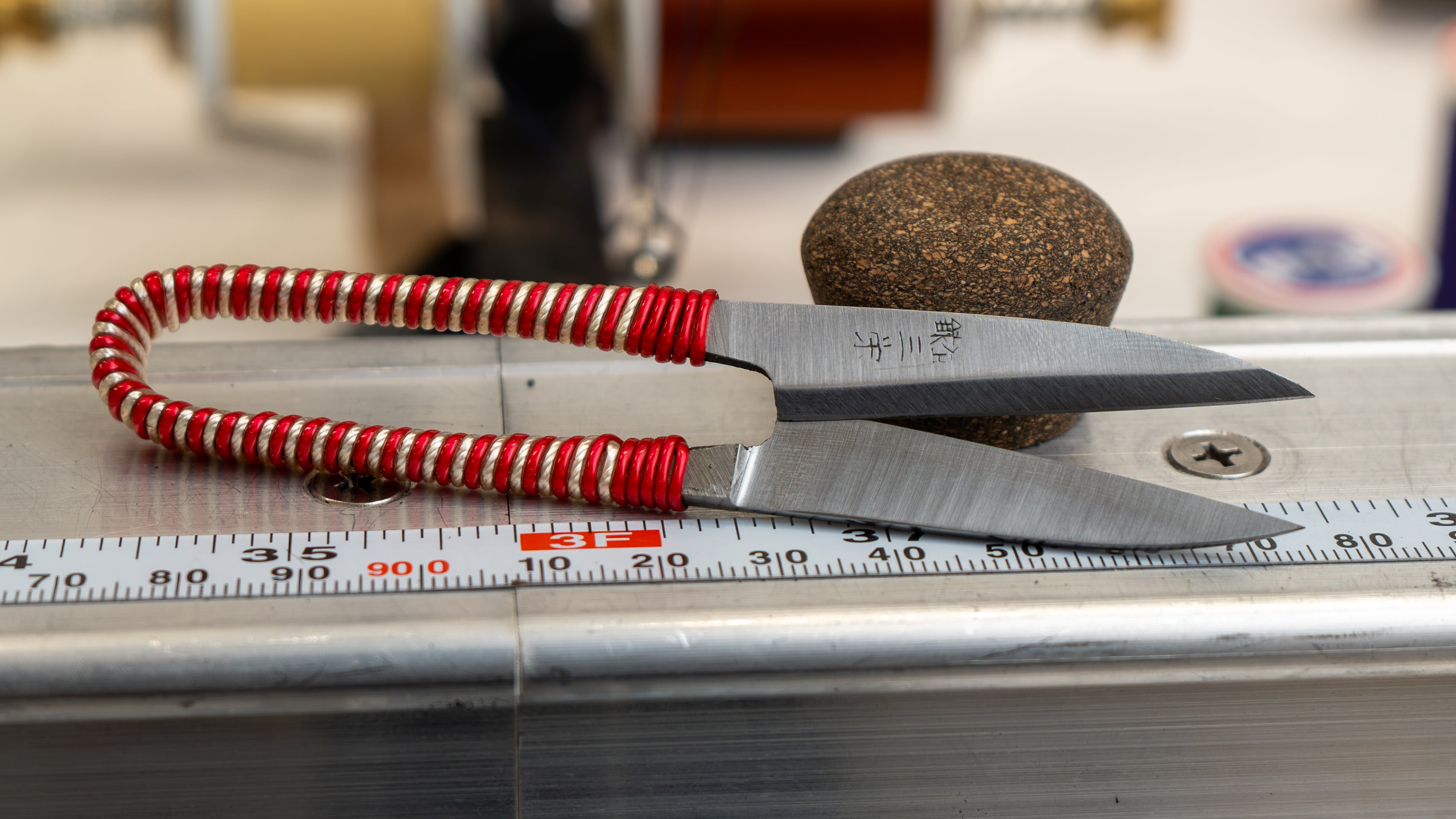 A close-up of a small scissors-style tool with a red-and-white striped handle and silver metal blades, resting on a metal surface with a measuring ruler and a round brown object in the background.